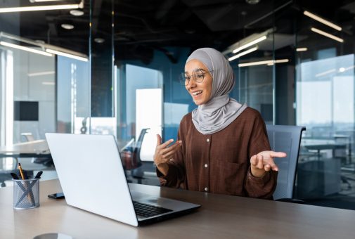 Successful and happy woman inside office at workplace, businesswoman boss in hijab talking on video call using laptop sitting at table muslim woman joyfully gesturing to colleagues in online meeting